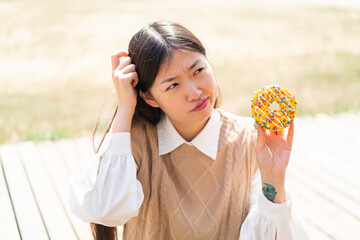 Young Chinese woman holding a donut at outdoors having doubts and with confuse face expression