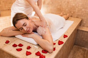 Beautiful woman enjoying foam massage in Turkish bath during attendant washes woman skin with foam....