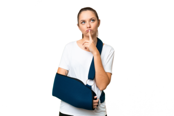 Young blonde woman with broken arm and wearing a sling over isolated chroma key background having doubts while looking up