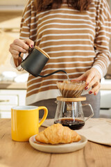 female makes coffee in morning kitchen. brews in purover. yellow cup with black coffee drip. daily routine of happy woman. Food and drinks, croissant on table. Americano cappuccino for cheerfulness