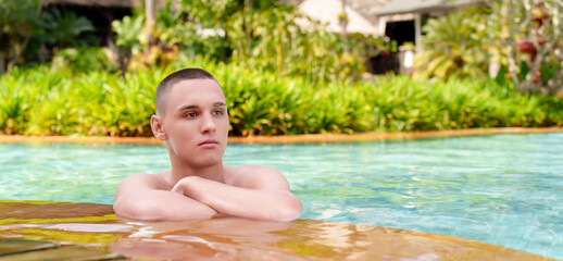A young guy at the edge of the pool enjoys the cool blue water of the hot summer. In the background there is a green planting of flowers.