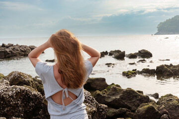The view from the back of a young girl with flowing long hair looks into the distance of the sea. Traveling alone, enjoying silence and peace, filling yourself.