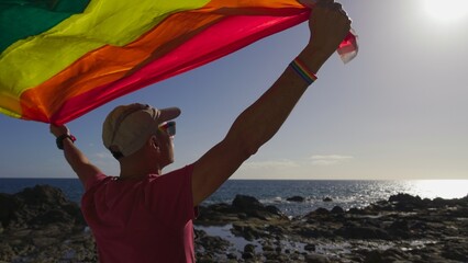 Young adult gay male proudly holds up the rainbow flag waving in the sun