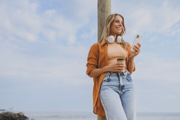 Bottom view young woman wear orange shirt casual clothes headphones listen music use mobile cell phone drink coffee walk on sea ocean sand shore beach outdoor seaside in summer day. Lifestyle concept.