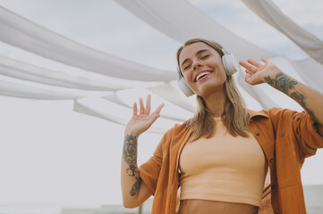 Bottom view young cheerful woman wears orange shirt beige top casual clothes listen to music in headphones raise up hands dance rest relax walk outside in summer day. Urban lifestyle leisure concept.