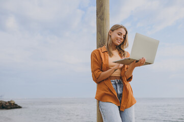 Side view smiling cheerful young IT woman she wears orange shirt casual clothes hold use work on laptop pc computer walk on sea ocean sand shore beach outdoor seaside in summer day. Lifestyle concept.