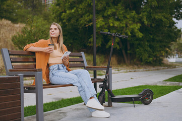 Full body side view young woman in shirt casual clothes use mobile cell phone drink coffee sit on bench near e-scooter walk rest relax in spring city park outdoors on nature. Urban lifestyle concept