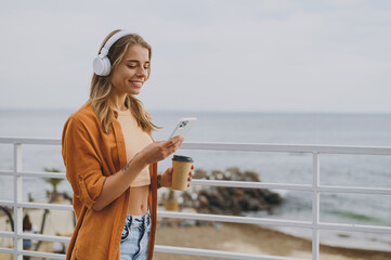 Side view young woman wears orange shirt casual clothes headphones listen music use mobile cell phone drink coffee walk on sea ocean sand shore beach outdoor seaside in summer day. Lifestyle concept.