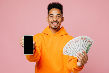 Young man of African American ethnicity he wearing yellow hoody casual clothes use blank screen mobile cell phone hold in hand fan of cash money in dollar banknotes isolated on plain pink background.