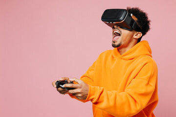 Side view young man of African American ethnicity in yellow hoody casual clothes hold in hand play pc game with joystick console watch in vr headset pc gadget isolated on plain light pink background