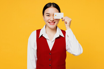 Young corporate lawyer employee business woman of Asian ethnicity wear formal red vest shirt work at office cover eye hold credit bank card isolated on plain yellow background studio. Career concept.