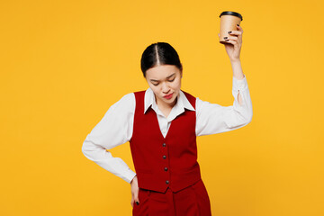 Young tired sad corporate lawyer employee business woman of Asian ethnicity wear formal red vest shirt work at office hold takeaway cup coffee to go isolated on plain yellow background Career concept