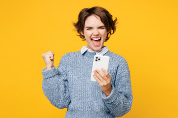 Young overjoyed woman she wears grey knitted sweater shirt casual clothes hold in hand use mobile cell phone do winner gesture isolated on plain yellow background studio portrait. Lifestyle concept.