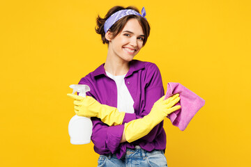 Young happy smiling woman she wears purple shirt rubber gloves do housework tidy up hold in hand rag spray bottle look camera isolated on plain yellow background studio portrait. Housekeeping concept. - obrazy, fototapety, plakaty