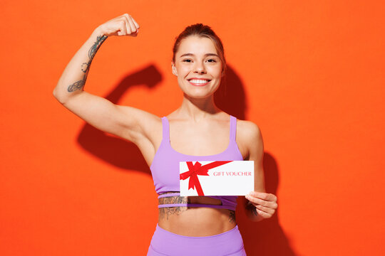 Young fitness trainer sporty woman sportsman wear purple top clothes in home gym hold gift coupon voucher card for store show muscles isolated on plain orange background Workout sport fit abs concept - Powered by Adobe