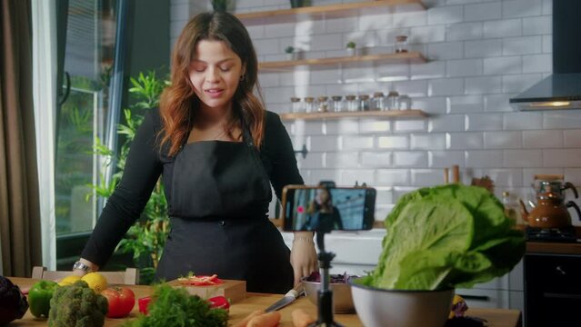 Young woman in an apron standing in kitchen records on smartphone new food videoblog. Woman cutting vegetables on a cutting board, blogging, healthy lifestyle, cooking master class event concept