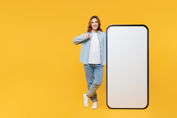 Full body young woman wears blue shirt white t-shirt casual clothes big huge blank screen mobile cell phone smartphone with area show thumb up isolated on plain yellow background. Lifestyle concept.