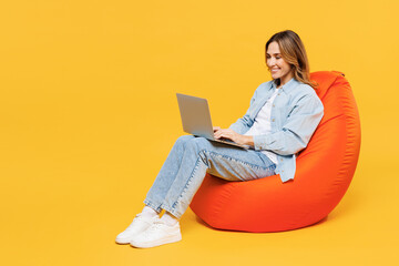 Full body young IT woman she wear blue shirt white t-shirt casual clothes sit in bag chair hold use work on laptop pc computer isolated on plain yellow background studio portrait. Lifestyle concept.