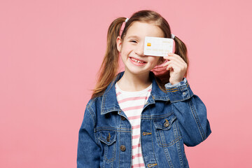 Little child cute kid girl 7-8 years old wears denim shirt hold in hand cover eye with mock up of credit bank card isolated on plain pastel pink background. Mother's Day love family lifestyle concept.