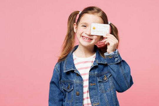 Little child cute kid girl 7-8 years old wears denim shirt hold in hand cover eye with mock up of credit bank card isolated on plain pastel pink background. Mother's Day love family lifestyle concept.