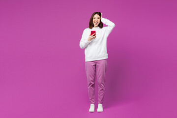 Full body young surprised excited happy woman wear light hoody casual clothes hold head use mobile cell phone look camera isolated on plain purple color background studio portrait. Lifestyle concept.