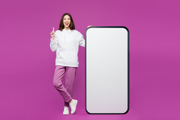 Full body young woman wear light hoody casual clothes big huge blank screen mobile cell phone smartphone with area point index finger up isolated on plain purple color background. Lifestyle concept.