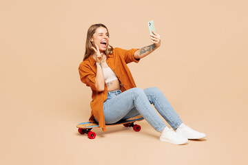 Full body young woman wear orange shirt casual clothes sits on pennyboard skateboard do selfie shot on mobile cell phone show v-sign isolated on plain pastel light beige background. Lifestyle concept.