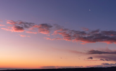 Fototapeta na wymiar La Luna all’alba resta ancora nel cielo sopra le nuvole rosa sulla colline mentre il sole sorge dal mare