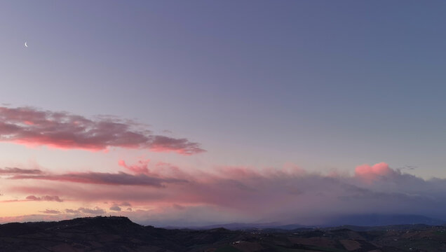 La Luna all’alba resta ancora nel cielo sopra le nuvole rosa sulla colline mentre il sole sorge dal mare © GjGj
