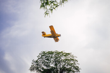 A vintage yellow biplane flying in the sky low above the ground sprays pesticides against insects....