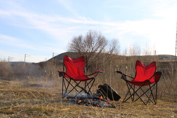 Two red chairs sit in front of a campfire with a blue sky in the background