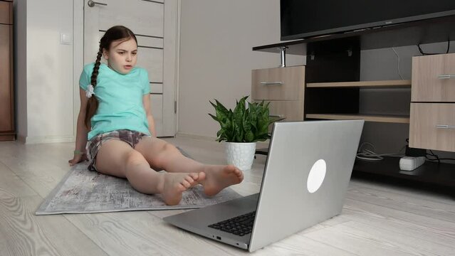 A little girl in homemade clothes is sitting on the floor in front of a laptop and doing an online workout, kneading the joints of her feet, lifting her leg up.