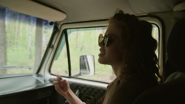 Happy girl with ginger long wavy hair sitting in front seat of camper van and talking to driver or her boyfriend on their way to countryside on summer weekendHappy girl with ginger long wavy hair sitt