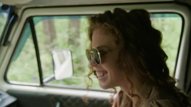 Happy young woman in sunglasses waving hand and laughing while sitting in camper van and chatting to fellow traveler on their way to countryside on summer weekend