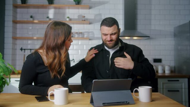 Young couple sit in kitchen while man suffering chest pain, holding massaging his chest, his girlfriend get worry and check him. Health problems concept