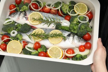 Woman putting baking dish with raw fish and vegetables into oven, top view
