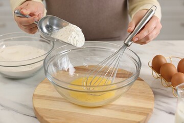 Woman whisking eggs while adding flour into bowl at light marble table indoors, closeup