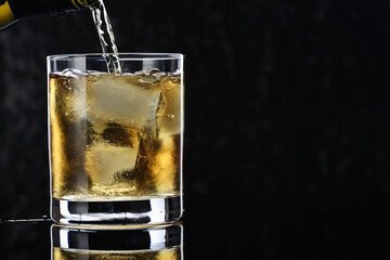 Pouring tasty whiskey from bottle into glass at mirror table against black background, closeup. Space for text