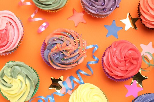 Delicious cupcakes with bright cream and party decor on orange background, flat lay