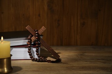 Bible, cross, rosary beads and church candle on wooden table, space for text