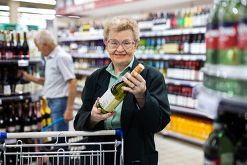 Mature woman with glasses chooses bottle of wine in alcohol section of supermarket