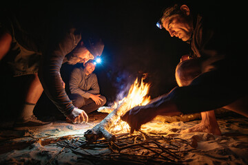 Three men gathered around a small campfire on a beach at night, two with headlamps