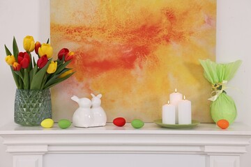 Easter decorations. Bouquet of tulips in vase, burning candles and bunny figures on fireplace at home