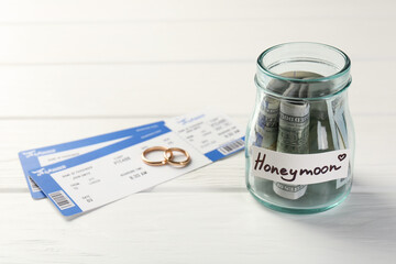 Glass jar with word Honeymoon, dollar banknotes, plane tickets and golden rings on white wooden table, closeup