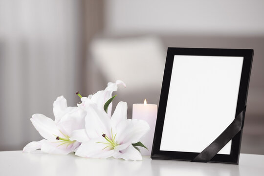 Funeral photo frame with black ribbon, white and violet lilies on white table indoors. Space for text
