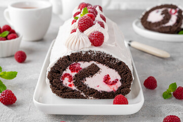 Chocolate roll cake with cream and fresh raspberries on a white plate on a gray background. Delicious holiday dessert.