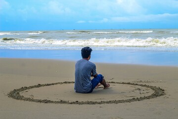 A sad man sitting down in sand looking at the sea. A young man sitting on a white sand beach during sunrise contemplating the scenery and the blue sea waves during his vacation in an idyllic nature.