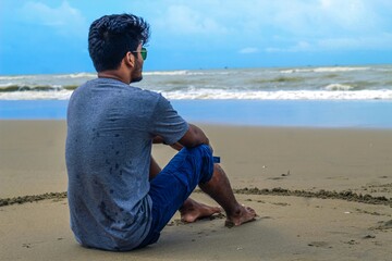 A sad man sitting down in sand looking at the sea. A young man sitting on a white sand beach during sunrise contemplating the scenery and the blue sea waves during his vacation in an idyllic nature.