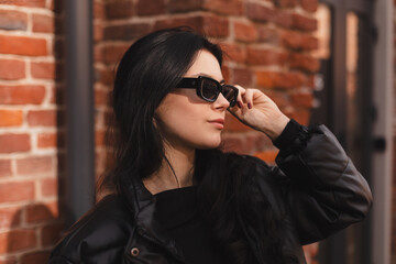 Pensive brunette woman in black puffer jacket put off black glasses and posing on office building...