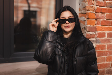 Pensive brunette woman in black puffer jacket put off black glasses and posing on office building...
